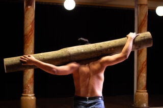 Eine hemdsärmelige Person steht in einem Haus und hält mit beiden Armen einen großen Ast über die Schultern.