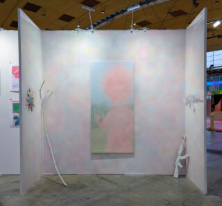 Eine pastellfarbene Kunstinstallation mit einem unscharfen Porträt in der Mitte, einem schwachen Text darunter, an der Wand befestigten Objekten und subtilen Regenbogenfarben auf drei weißen Tafeln in einem Galerieraum.