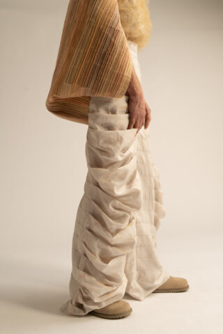 Eine Person, die eine cremefarbene Rüschenhose, beigefarbene Schuhe und einen strukturierten hellbraunen Ärmel trägt, steht vor einem schlichten Hintergrund.