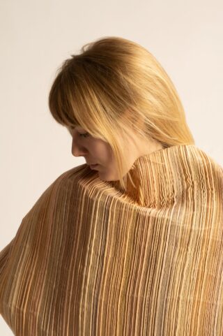 Eine Frau mit glattem blondem Haar blickt über ihre Schulter und trägt ein strukturiertes Kleidungsstück mit vertikalen beigen und braunen Streifen vor einem neutralen Hintergrund.