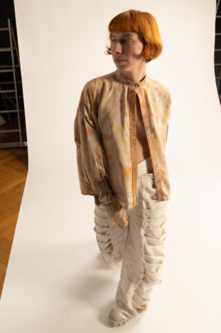 Eine Person mit kurzen orangefarbenen Haaren, die eine lockere, gemusterte Jacke und eine strukturierte weiße Hose trägt, steht in einem Studio mit weißem Hintergrund und schaut zur Seite.