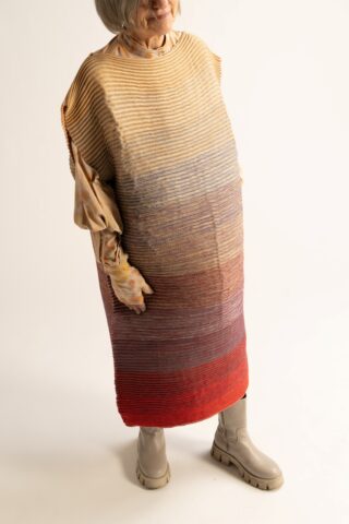 Eine ältere Frau in einem lockeren, gestreiften Kleid mit Farbverlauf in Gelb, Blau und Rot, langen Ärmeln und beigen Stiefeln steht vor einem schlichten Hintergrund.