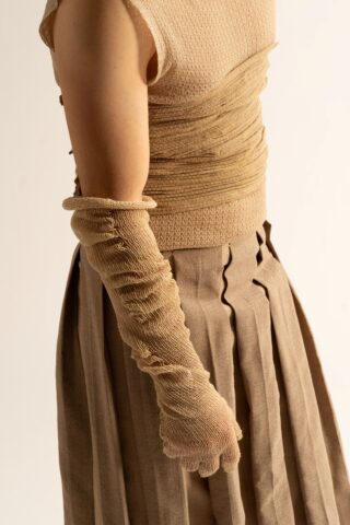 Eine Person, die ein ärmelloses beigefarbenes Oberteil, lange beigefarbene strukturierte Handschuhe und einen taupefarbenen Faltenrock trägt, fotografiert von der Seite vor einem unifarbenen Hintergrund.