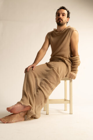 Eine Person in einem ärmellosen, strukturierten beigen Kleid sitzt seitwärts auf einem Holzhocker vor einem schlichten, hellen Hintergrund.