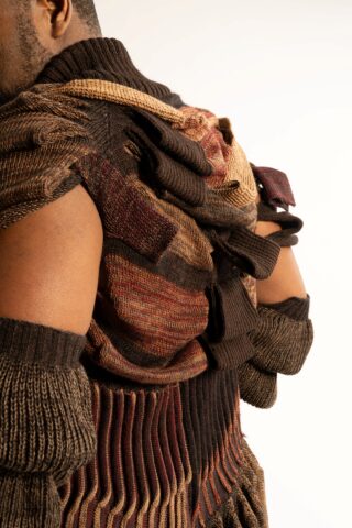 Eine Person, die ein strukturiertes, mehrfarbiges Strickkleidungsstück mit Lagenmustern in Braun-, Kastanien- und Brauntönen trägt, von hinten und von der Seite gesehen.