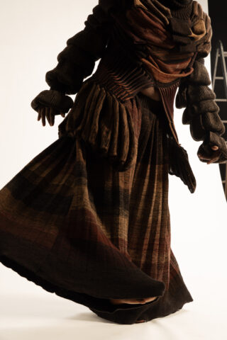 Eine Person, die ein strukturiertes, mehrlagiges braunes Outfit mit voluminösen Ärmeln und einem langen, karierten Rock trägt, steht vor einem schlichten Hintergrund.