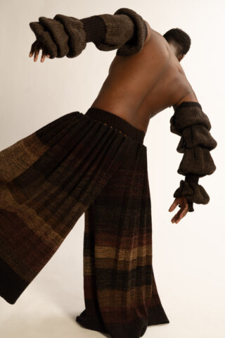 Eine Person, die eine strukturierte, erdfarbene Strickhose und passende Armstulpen trägt, posiert mit entblößtem Rücken vor einem schlichten Hintergrund.