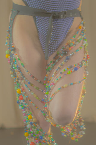 Eine Person, die einen strukturierten Body und einen Gürtel mit mehrfarbigen Perlensträngen trägt, die über ihre Oberschenkel drapiert sind.