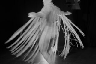 In einem dunklen Raum mit Bühnenbeleuchtung dreht sich eine Person mit ausgestreckten Armen, die ein Kleidungsstück mit langen, fließenden Fransensträngen trägt.