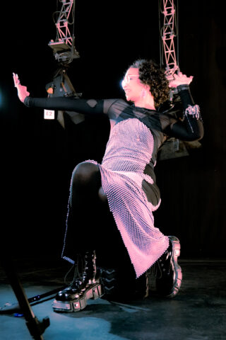 Eine Person, die ein schwarzes Netz-Outfit mit silbernen Verzierungen und Plateaustiefeln trägt, kniet auf einem Knie und posiert mit einem erhobenen Arm im Bühnenlicht.