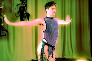 Eine Person in einem ärmellosen, mit Pailletten besetzten Netz-Outfit steht mit ausgestreckten Armen auf der Bühne, die von grünem und gelbem Licht beleuchtet wird, während im Hintergrund Vorhänge und Bühnenscheinwerfer zu sehen sind.