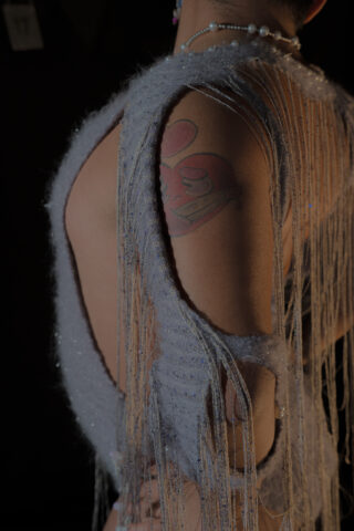 Eine Person, die ein graues, strukturiertes, nach hinten offenes Kleidungsstück mit Fransen trägt, das eine große rote Tätowierung auf dem Oberarm zeigt.