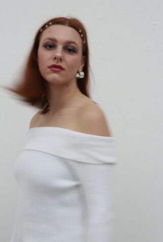 Eine Frau mit roten Haaren, die ein weißes schulterfreies Oberteil und Perlenohrringe trägt, steht vor einem schlichten weißen Hintergrund, der teilweise verschwimmt, wenn sie ihren Kopf dreht.