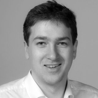 Ein Mann mit kurzem, dunklem Haar lächelt in die Kamera. Er trägt ein helles Hemd mit Kragen. Der Hintergrund ist schlicht und neutral.