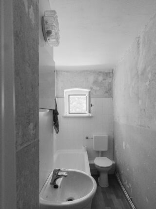 Kleines, schmales Badezimmer mit Waschbecken, Badewanne, Toilette, unten gefliesten Wänden, oben abblätternden Wänden und einem kleinen Fenster, das Tageslicht hereinlässt.