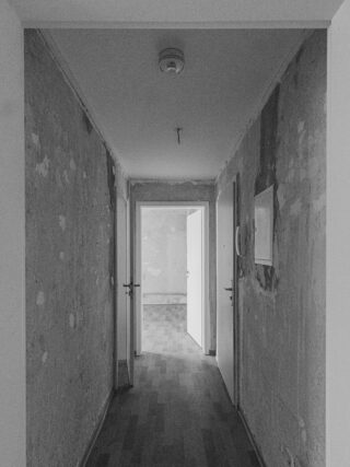 Ein schmaler, schwach beleuchteter Flur mit abgenutzten, abblätternden Wänden führt zu einem Raum mit offenen weißen Türen und einem Holzboden.