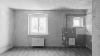 Leeres, unfertiges Zimmer mit kahlen Wänden, zwei Fenstern, zwei Heizkörpern und einem Teil der freiliegenden Rohrleitungen. Der Boden ist mit Holzlaminat ausgelegt. Es sind keine Möbel oder Dekorationen vorhanden.