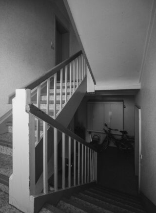 Schwarz-Weiß-Foto einer Innentreppe mit Geländer, einer geschlossenen Tür am Treppenabsatz und zwei Fahrrädern, die in der Nähe eines Schranks unter der Treppe abgestellt sind.