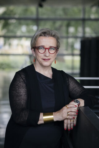 Eine Frau mit kurzen grauen Haaren, roter Brille und Goldschmuck steht in einem Innenraum, trägt ein schwarzes Outfit und schaut mit neutralem Blick in die Kamera.