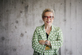 Eine Frau mit kurzen grauen Haaren, roter Brille und grün gemusterter Jacke steht lächelnd mit verschränkten Armen vor einer Betonwand.