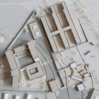 Architekturmodell mit rechteckigen und quadratischen Gebäuden, die auf einem konturierten Sockel angeordnet sind, mit Wegen, kleinen Strukturen und minimalen Landschaftselementen.