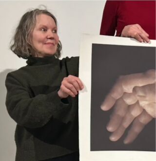 Eine Frau hält ein großes Foto mit sich überlappenden Händen in der Hand, während eine andere Person, die teilweise sichtbar ist, bei der Darstellung des Bildes hilft.