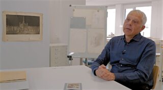 Ein Mann in einem blauen Hemd sitzt an einem Schreibtisch in einem Büro, mit Dokumenten und einem Zeichenbrett im Hintergrund.