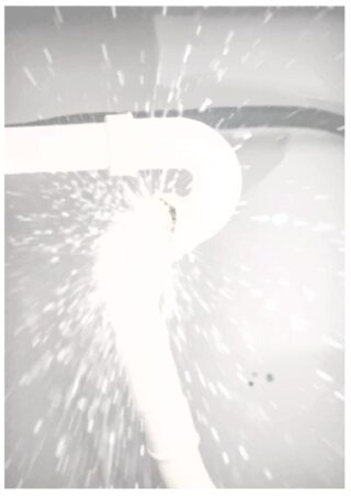 Wasser, das aus einem Leck in einer weißen PVC-Rohrverbindung vor einem grauen Hintergrund spritzt.