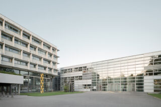 Modernes mehrstöckiges Gebäude mit großen Glasfenstern, horizontalen Linien und einer gelben vertikalen Skulptur im offenen Innenhof unter freiem Himmel.