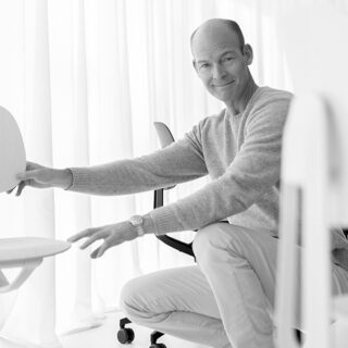 Ein Mann in einem hellen Pullover kniet in einem hellen, minimalistischen Raum mit durchsichtigen Vorhängen und verstellt einen Stuhl.