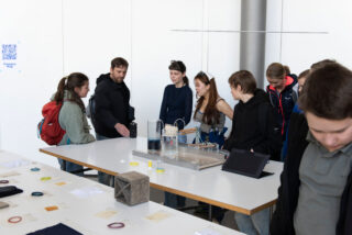 Eine Gruppe von Personen steht in einem gut beleuchteten Raum um einen Demonstrationstisch mit wissenschaftlichen Geräten und Proben herum und beobachtet und bespricht die Ausstellung.