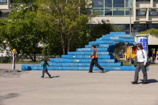 Drei Personen gehen an einer großen blauen Kunstinstallation aus gestapelten Kisten vor einem modernen Gebäude vorbei, mit Bäumen im Hintergrund.