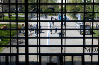 Blick durch große Glasfenster auf Menschen, die sich auf einem gepflasterten Gehweg im Freien versammelt haben, wobei einige in der Nähe eines langen Streifens auf dem Boden und andere bei Ständen oder Auslagen stehen.