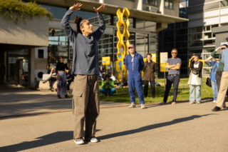 Eine Person steht im Freien mit erhobenen Armen, während eine Gruppe von Menschen im Hintergrund in der Nähe eines modernen Gebäudes und einer gelben Skulptur zusieht.