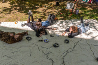 Vier Personen liegen auf einem großen weißen Laken im Freien, jede mit Kopfhörern und Audiogeräten, umgeben von Bäumen und gedämpftem Sonnenlicht.