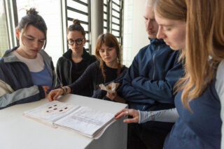 Fünf Personen betrachten aufmerksam ein aufgeschlagenes Buch oder Heft, das auf einem weißen Sockel in einem gut beleuchteten, modernen Innenraum ausgestellt ist.