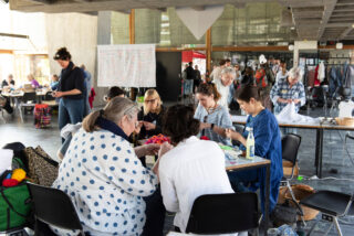 Eine Gruppe von Menschen sitzt um einen Tisch herum und arbeitet mit Textilien, während andere in einem großen, gut beleuchteten Raum mit Betonwänden und Fenstern stehen und sich bewegen.