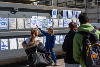 Eine Frau in Blau zeigt auf Plakate zum Tag der Erde und zu Klimafragen an einer Wand, während eine kleine Gruppe von Menschen davor steht und zuhört.