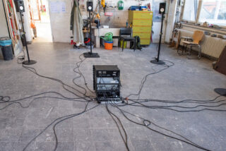 Auf dem Boden einer Industriehalle sind Audiogeräte mit mehreren Kabeln aufgestellt, die mit im Raum verteilten Lautsprechern verbunden sind.