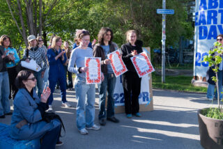 Eine Gruppe von Menschen steht im Freien und hält Urkunden in der Hand, während andere klatschen; auf einem Schild im Hintergrund steht 