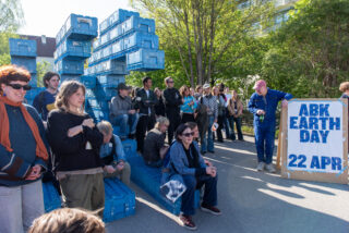 Eine Gruppe von Menschen versammelt sich im Freien neben gestapelten blauen Kisten und einem großen Schild mit der Aufschrift 
