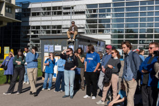 Eine Gruppe von Menschen steht vor einem modernen Gebäude, einige tragen blaue Hemden, während eine Person auf einem mit Plakaten bedeckten Metallcontainer sitzt.