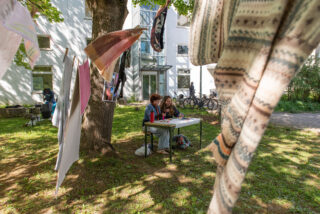 Zwei Personen sitzen an einem Tisch unter einem Baum und schreiben oder zeichnen, während in der Nähe gemusterte Stoffe hängen und ein weißes Gebäude im Hintergrund zu sehen ist.