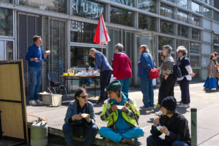 Die Menschen stehen in einer Schlange im Freien vor einem Gebäude mit Glasfenstern, während andere etwas essen; ein Tisch mit Vorräten und eine rote Fahne sind zu sehen.