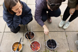 Drei Personen hocken auf dem Bürgersteig und betrachten drei Töpfe, die mit Wasser und Garn in Braun-, Rot- und dunklen Violetttönen gefüllt sind. Sie benutzen Holzstöcke, um den Inhalt umzurühren.