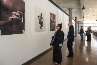 Mehrere Personen stehen in einer Galerie und betrachten große Fotografien von menschlichen Figuren und Körperteilen, die an einer weißen Wand angebracht sind.