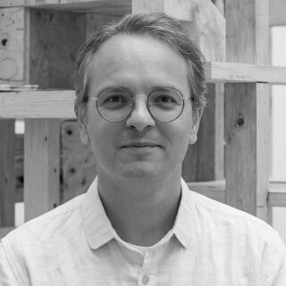 Eine Person mit kurzen hellen Haaren und einer runden Brille, die ein helles Hemd mit Knöpfen trägt, steht vor einem Holzbau.
