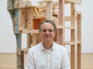 Ein Mann mit Brille und weißem Hemd steht in einer Galerie vor einer abstrakten Holzskulptur.