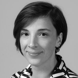 Eine Person mit kurzen dunklen Haaren, die ein gemustertes Hemd und kleine Ohrringe trägt, schaut in diesem Schwarz-Weiß-Porträt in die Kamera und lächelt leicht.