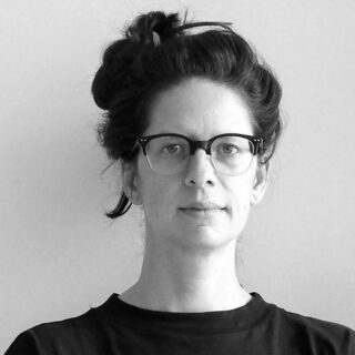 Eine Person mit dunklem Haar in einem unordentlichen Dutt, die eine Brille und ein schwarzes Hemd trägt, blickt vor einem schlichten hellen Hintergrund direkt in die Kamera.